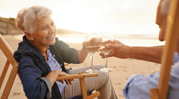 A happy senior couple on the beach smiling and toasting with wine glasses.