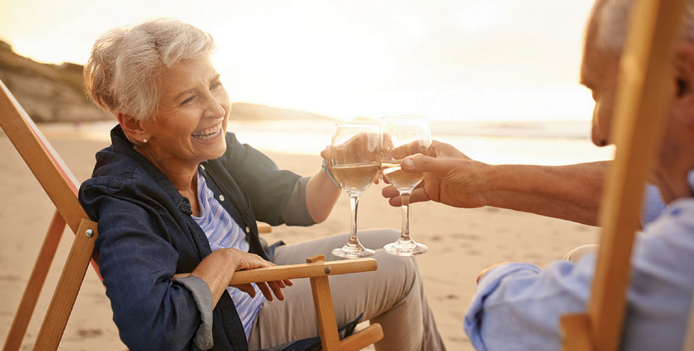 A happy senior couple on the beach smiling and toasting with wine glasses.